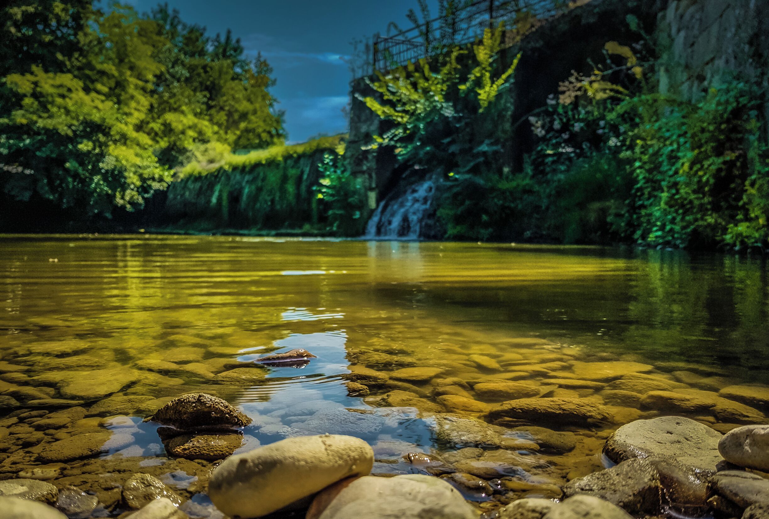 A clear river with rocks and a waterfall in the background.