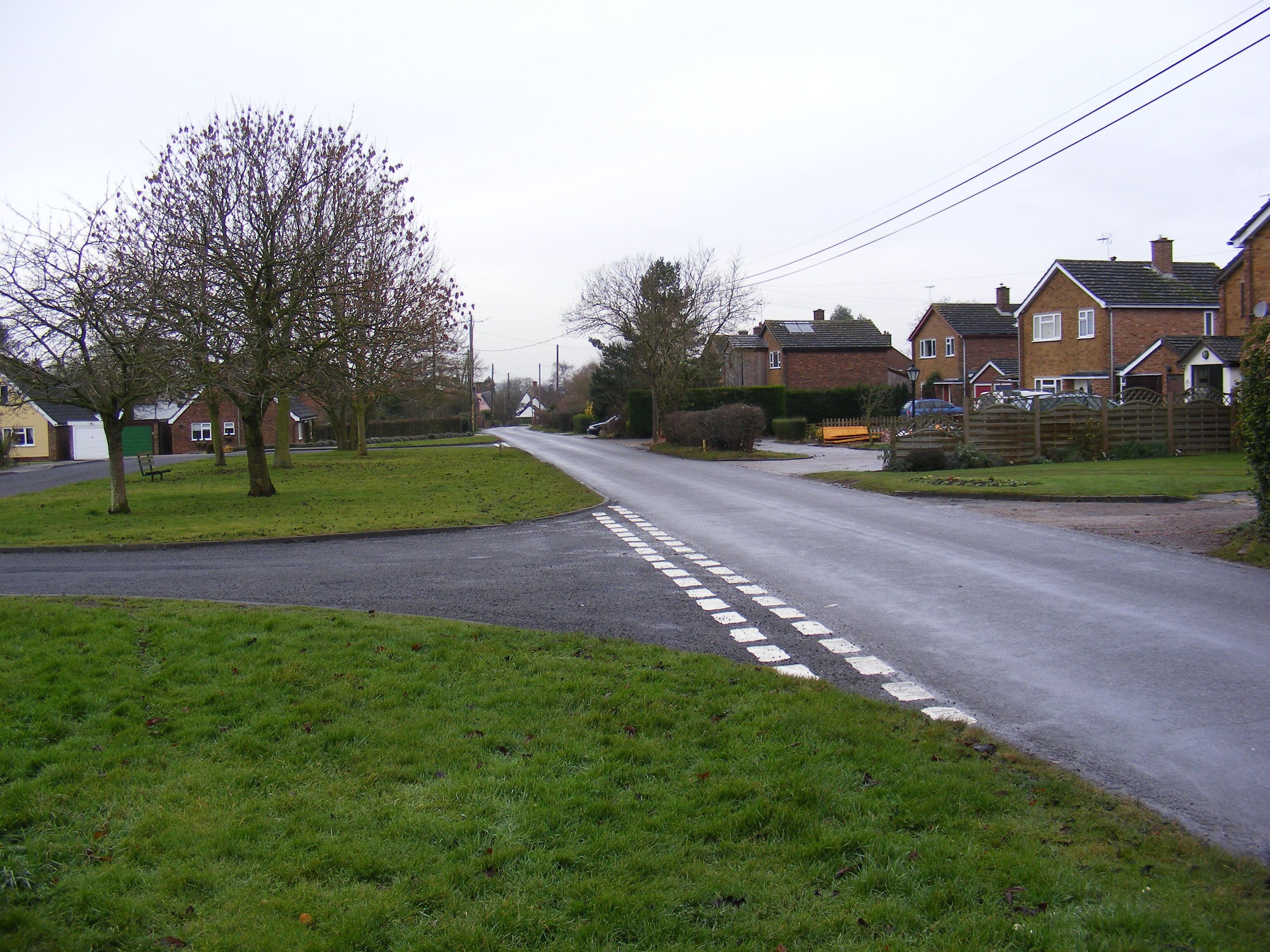 A quiet suburban street with houses, a grassy area, and a tree-lined road.