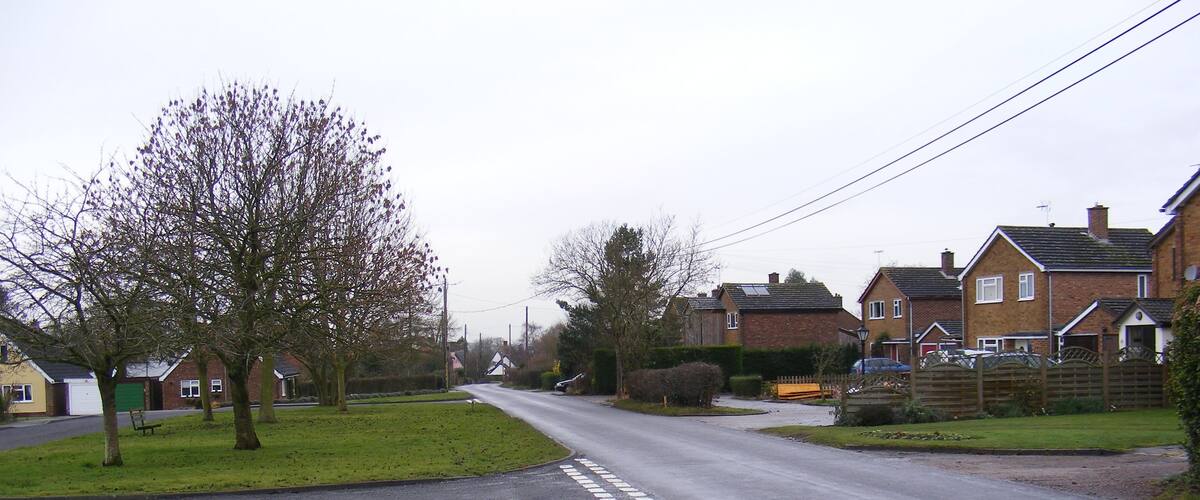 A quiet suburban street with houses, a grassy area, and a tree-lined road.