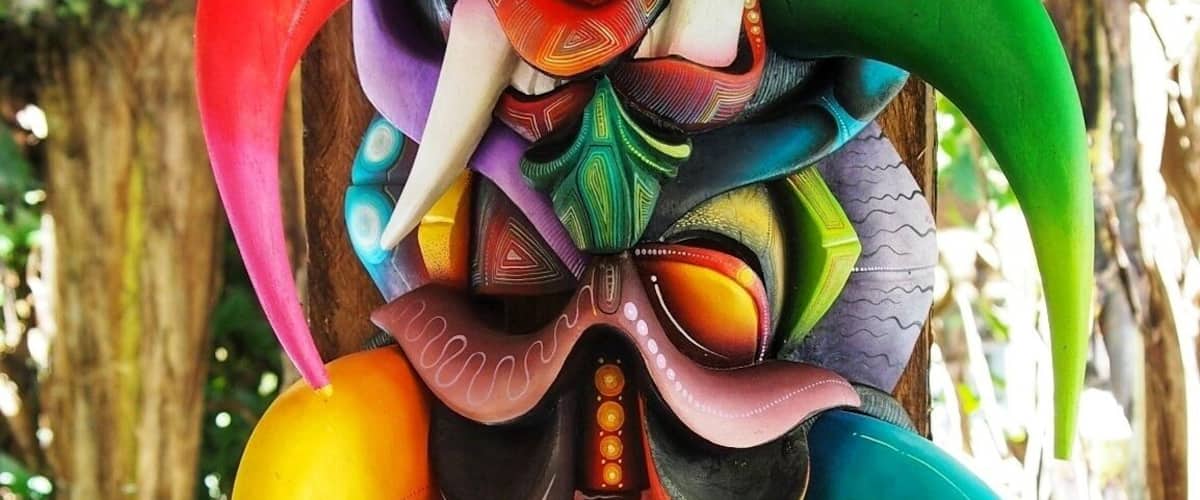 A colorful carved mask on a wooden post in a forest.