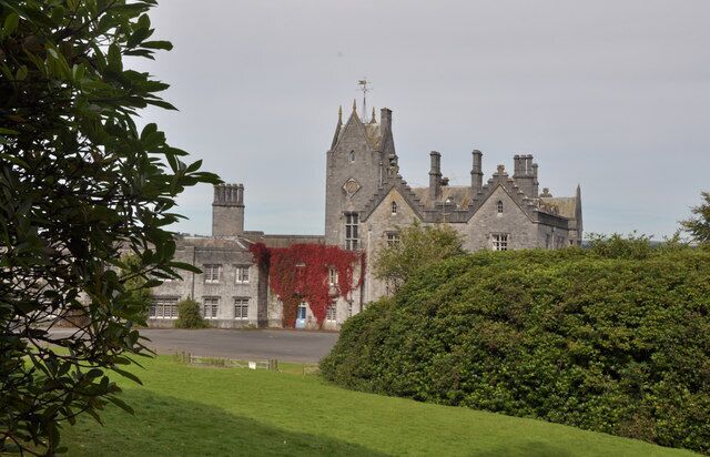 A large stone building with a tower, surrounded by green hedges and a grassy area.