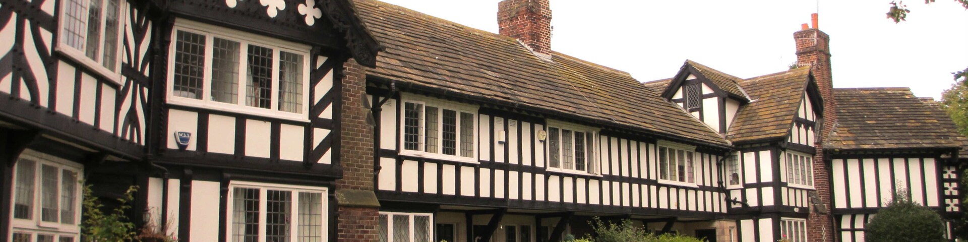 Um edifício em estilo Tudor com estrutura de madeira preta e branca, chaminés de tijolo e um jardim bem cuidado com sebes e gramado.