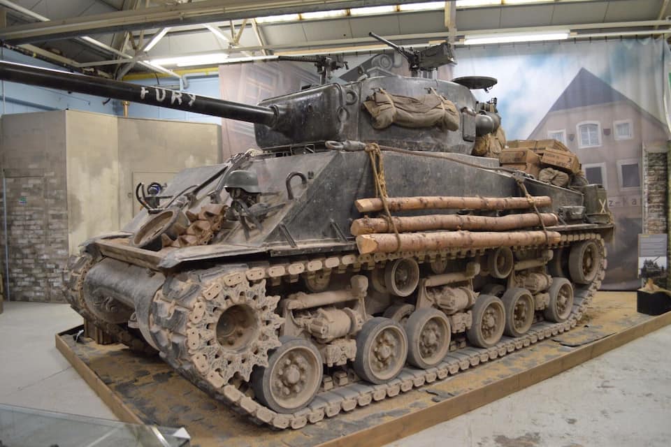 A military tank with a mounted gun and tracks, displayed indoors with a backdrop of a building and a banner.