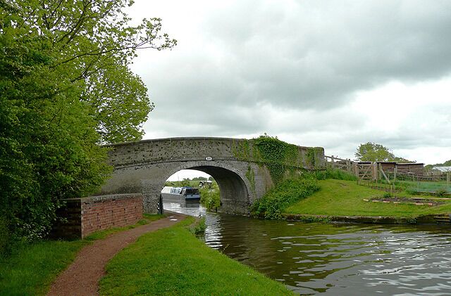 A stone bridge over a canal with a path beside it, surrounded by greenery and a cloudy sky.