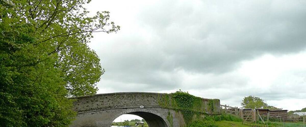 A stone bridge over a canal with a path beside it, surrounded by greenery and a cloudy sky.