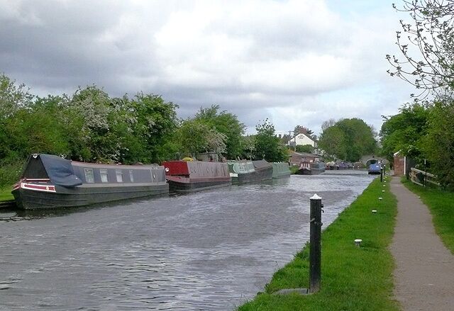 Shropshire Union Canal at Wheaton Aston, Staffordshire. The Shropshire Union Canal was opened in 1835 as the Birmingham and Liverpool Junction Canal, engineered by Thomas Telford. The picture shows the scene from Wheaton Aston Lock. The black pillar in the foreground is a water point for boats. The brick building further away by the tow-path is a boaters' sanitary station. The white building past the boats is The Hartley Arms, by Tavern Bridge.