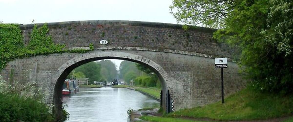 Wheaton Aston Bridge, Staffordshire. This is bridge No 18 on the Shropshire Union Canal, which was opened in 1835 as the Birmingham and Liverpool Junction Canal, engineered by Thomas Telford. Through the arch you will see Wheaton Aston Lock, the first one since the stop lock at Autherley eight miles to the south-east. Telford's policy was to create the shortest canal possible with his technology, building many cuttings and embankments to avoid the alternatives of increasing the distance by meandering along the contours (Brindley's 18th century style) and building locks, both of which would have slowed traffic considerably. Railways were, at that time, taking an increasing amount of the canal companies' business.