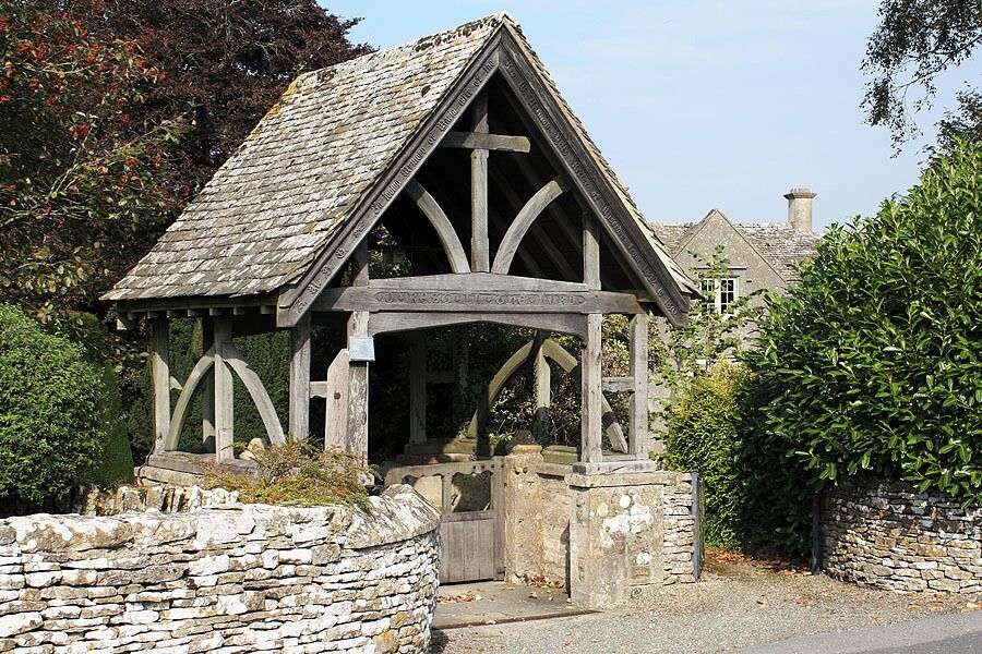 A stone archway with a wooden structure above it, surrounded by greenery and a stone wall.
