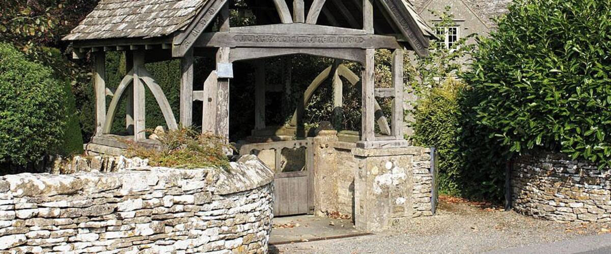 A stone archway with a wooden structure above it, surrounded by greenery and a stone wall.