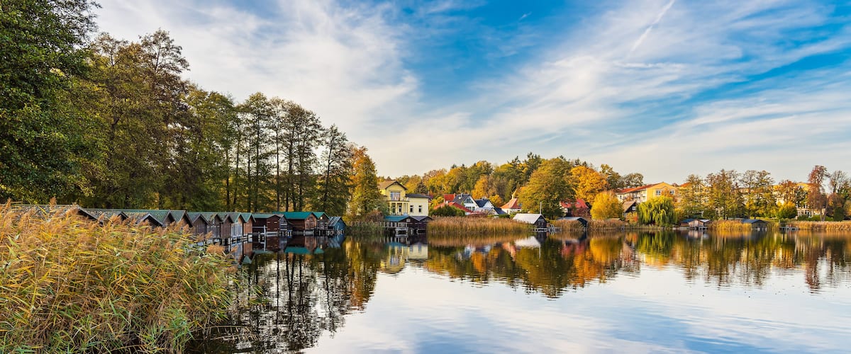 A calm lake with reflections of houses and trees, surrounded by a forest and a clear sky.