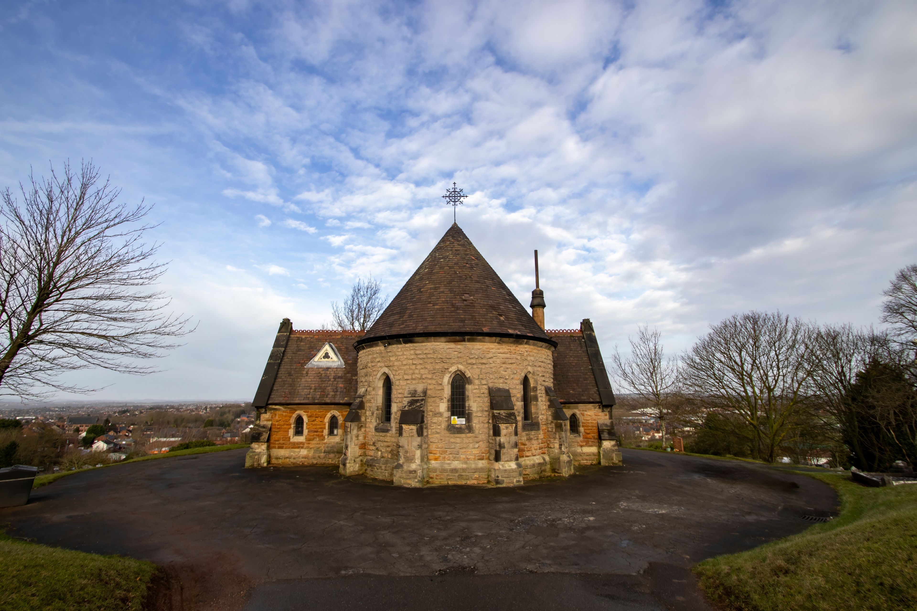 Une église historique en pierre, dotée d’un toit conique et d’une girouette, entourée d’une allée circulaire et d’arbres sans feuilles.