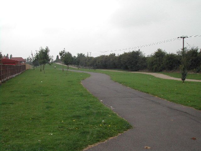 A paved path curving through a grassy area with trees and a fence on one side.
