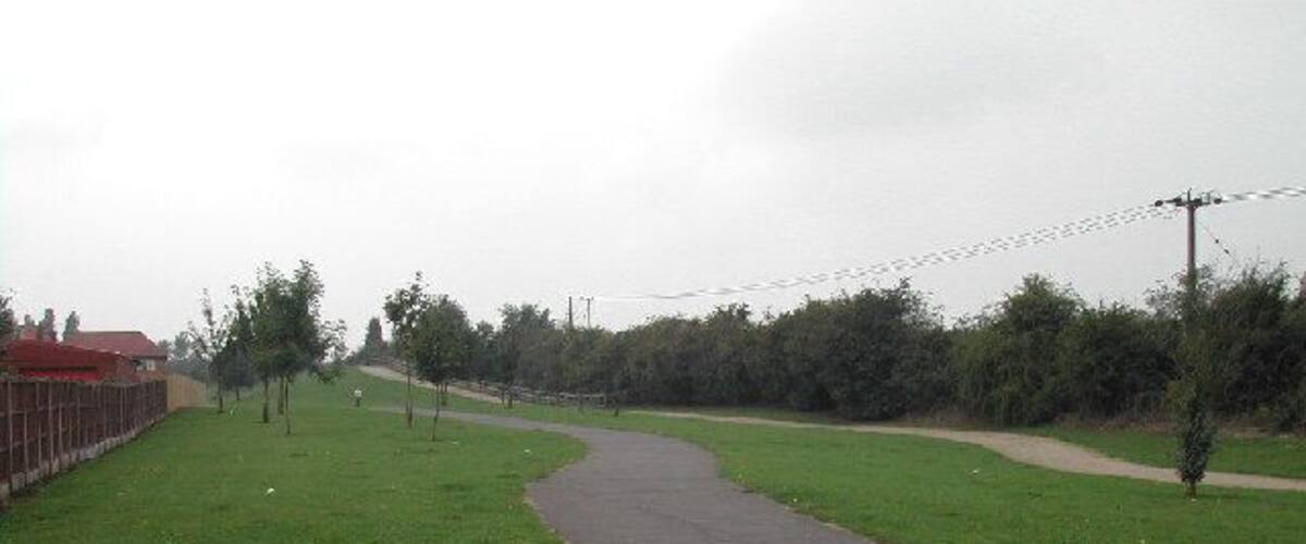 A paved path curving through a grassy area with trees and a fence on one side.