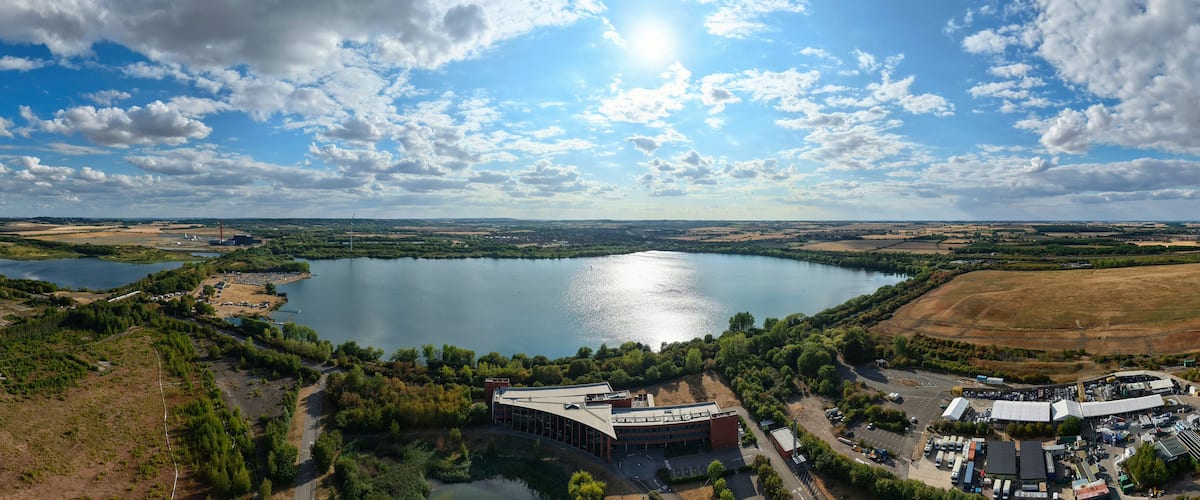 Un lago extenso rodeado de vegetación, un complejo de edificios y campos abiertos.