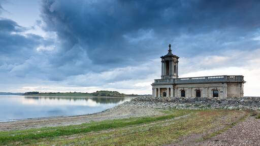 A historic stone building with a tower by a calm lake under a cloudy sky.