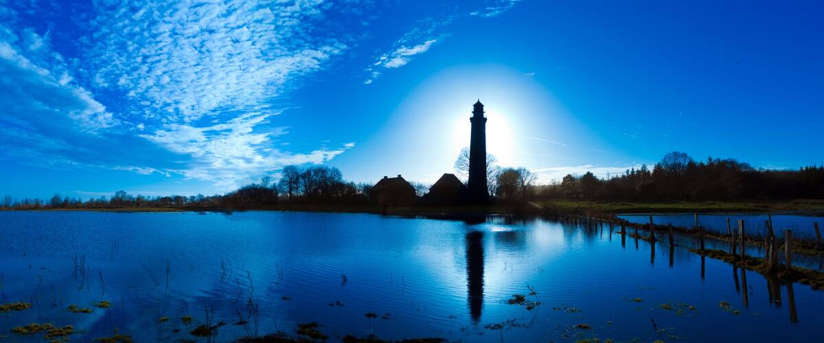 A calm lake with a lighthouse in the distance, surrounded by trees and a clear blue sky.