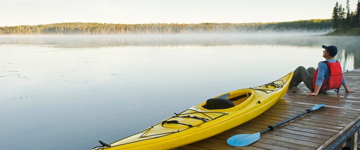 A person sitting on a dock next to a yellow kayak and a paddle.