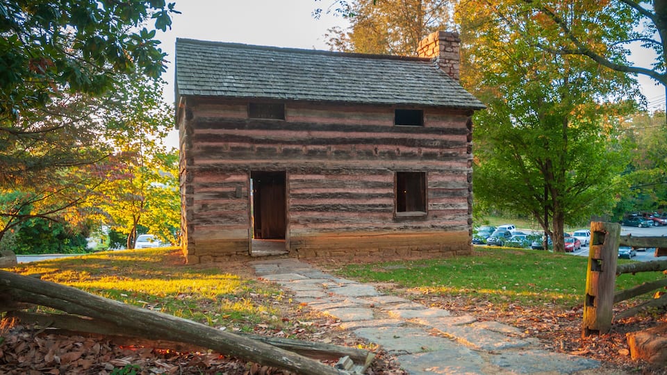 A rustic log cabin with a stone foundation, surrounded by autumn foliage and a stone pathway.