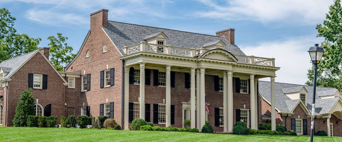 A large brick building with a covered porch and columns, surrounded by well-maintained lawns and trees.