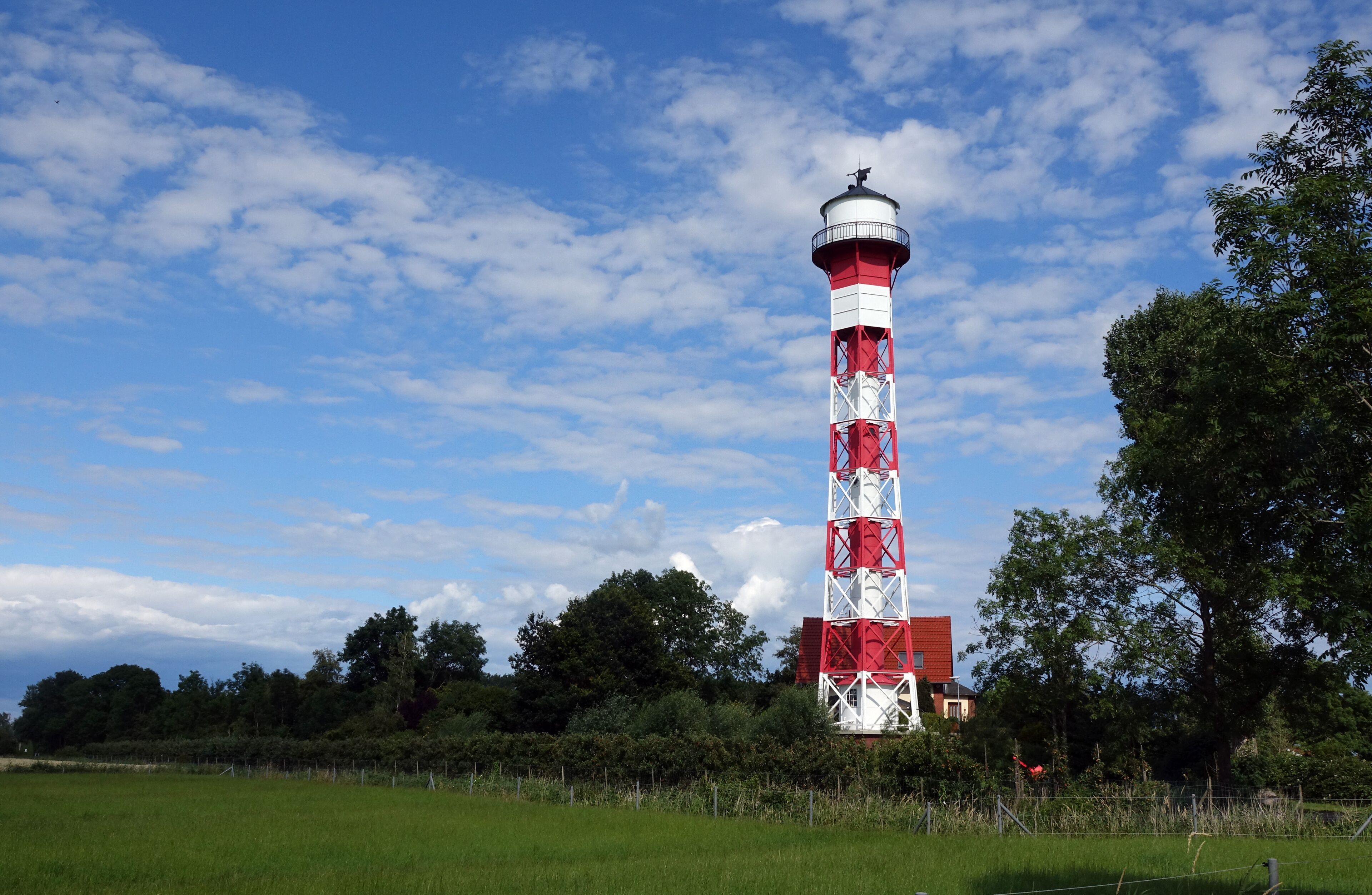 A red and white lighthouse in a grassy field with trees and a clear sky.