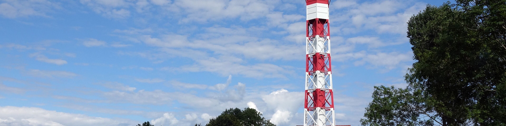 A red and white lighthouse in a grassy field with trees and a clear sky.