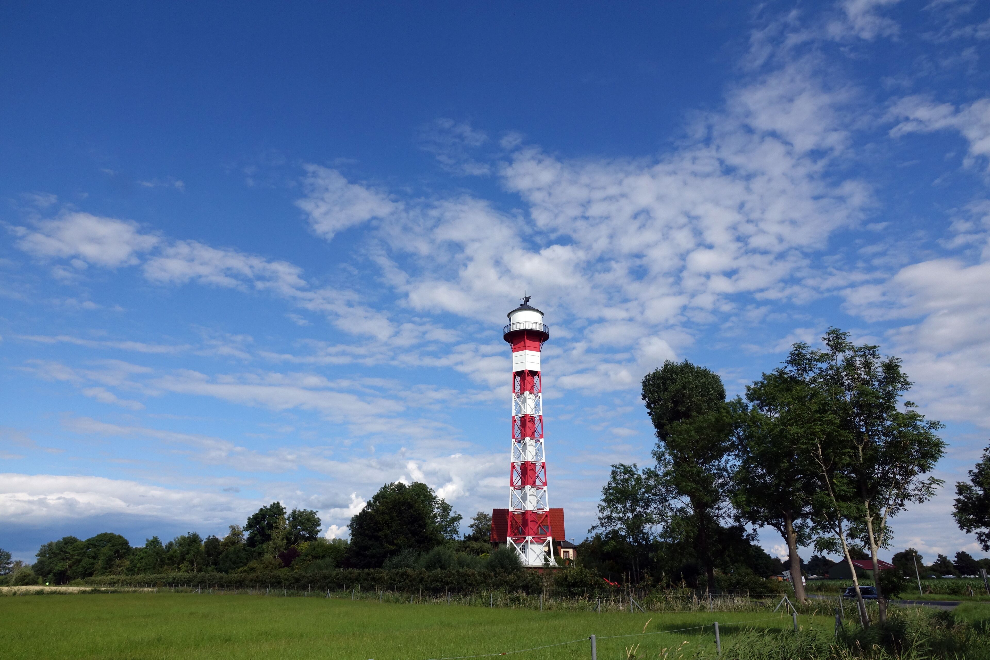 A red and white lighthouse in a grassy field with trees and a blue sky.