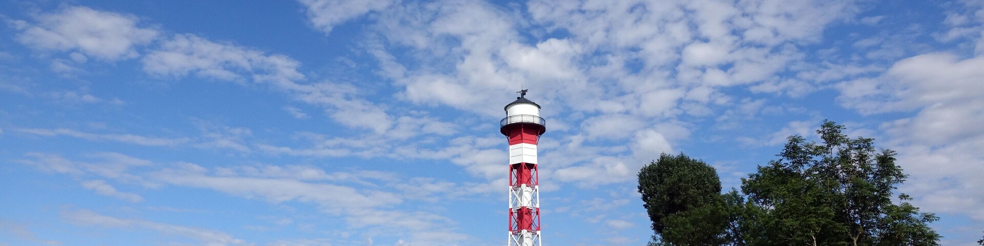 A red and white lighthouse in a grassy field with trees and a blue sky.