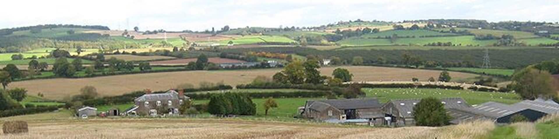 A rural landscape with fields, a farmhouse, and rolling hills in the background.