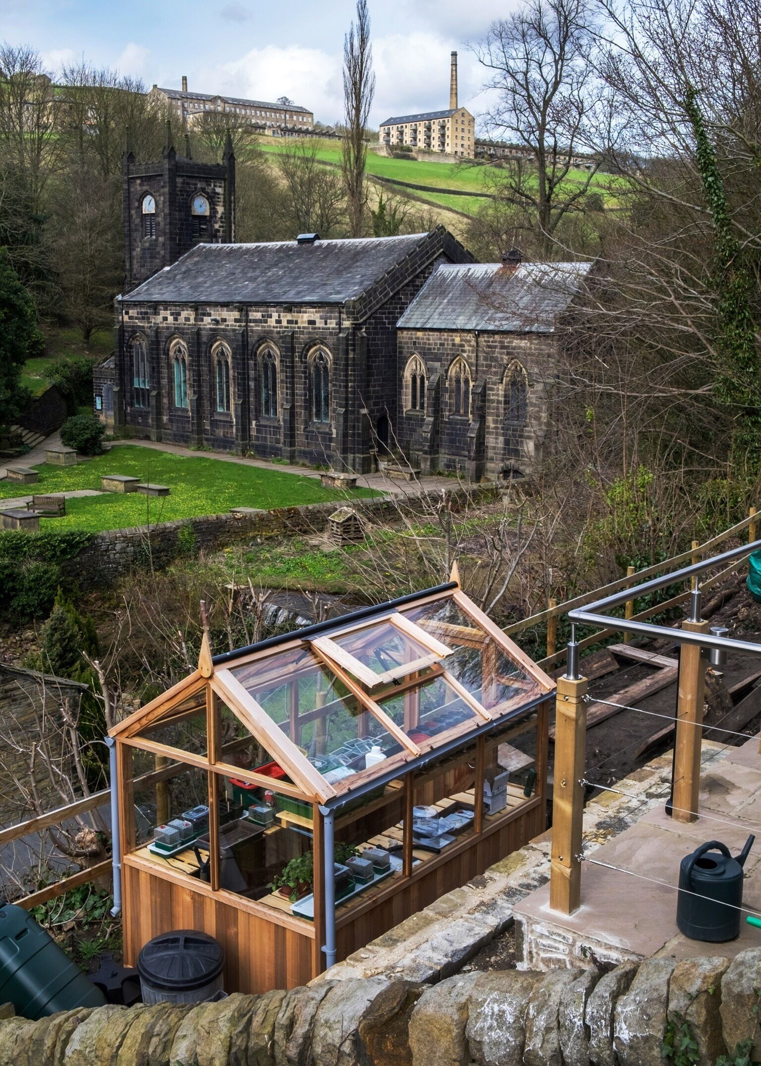 A historic stone church with a clock tower, surrounded by a green landscape and a modern glass greenhouse structure.
