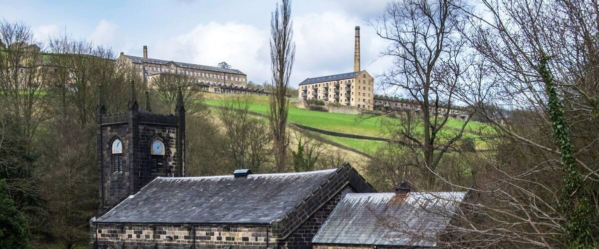 A historic stone church with a clock tower, surrounded by a green landscape and a modern glass greenhouse structure.