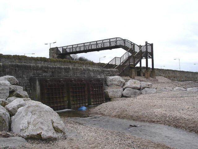 A stone bridge with a metal staircase leading up to it, surrounded by large rocks and a gravel path.