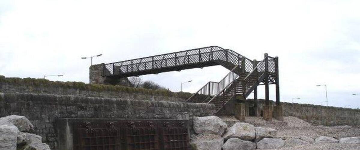 A stone bridge with a metal staircase leading up to it, surrounded by large rocks and a gravel path.