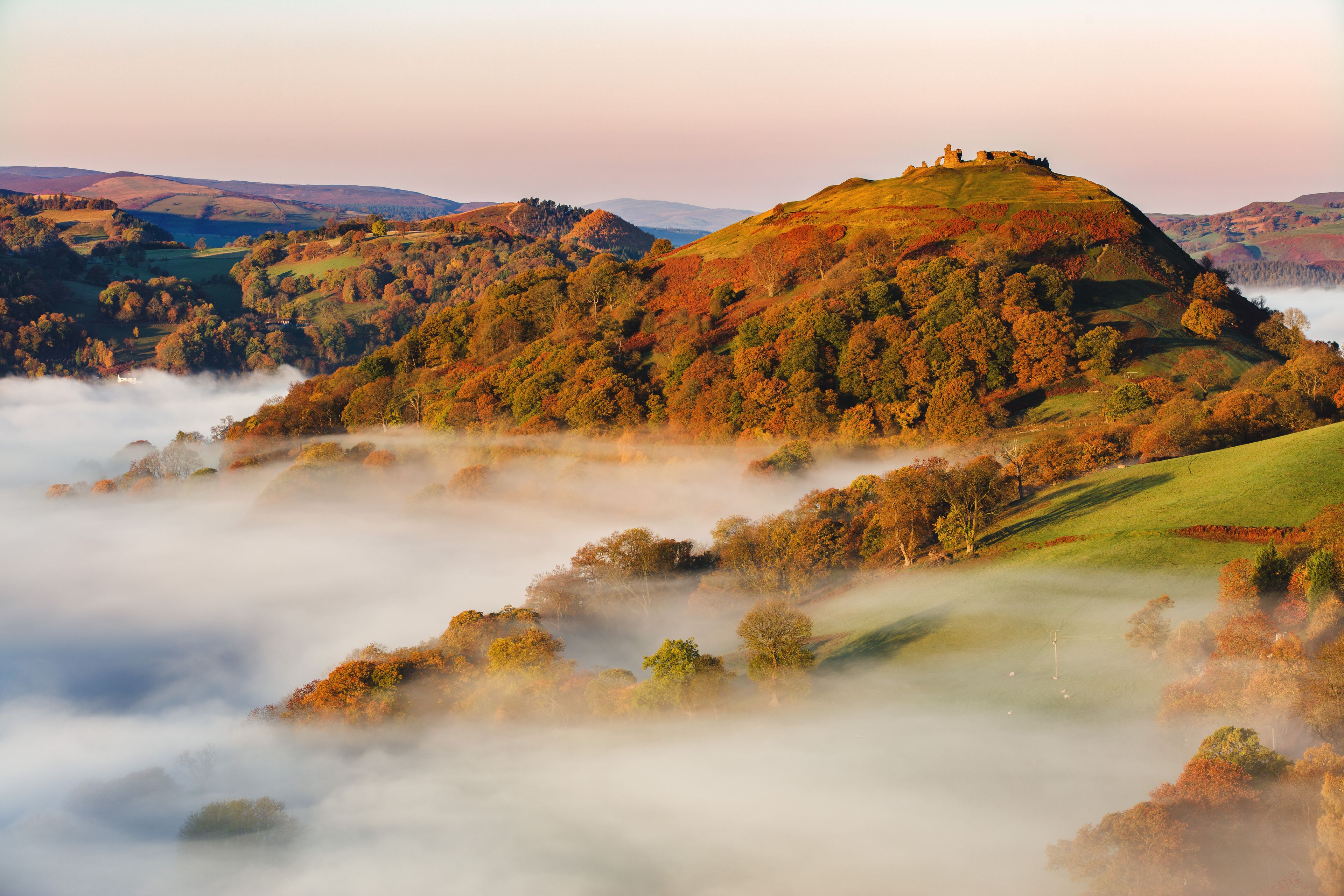A misty valley with rolling hills, a prominent hilltop with ruins, and a distant view of a lake.