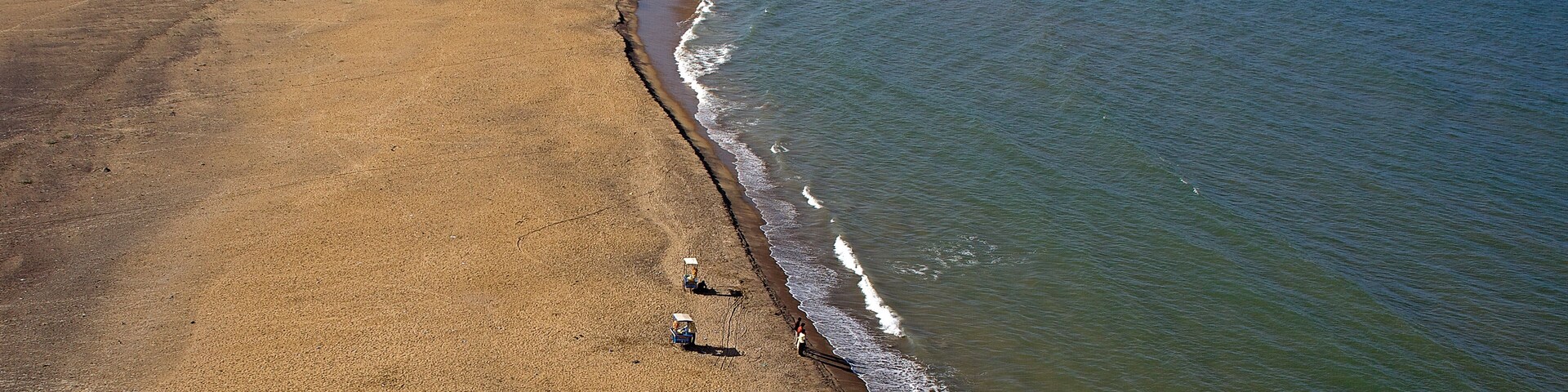 Vista aérea de uma praia com uma extensa faixa de areia, uma estreita faixa de vegetação e o oceano.