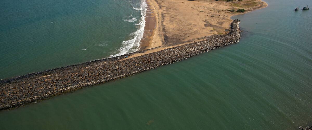 An aerial view of a coastal area with a sandy beach, a rocky breakwater, and a river flowing into the sea.