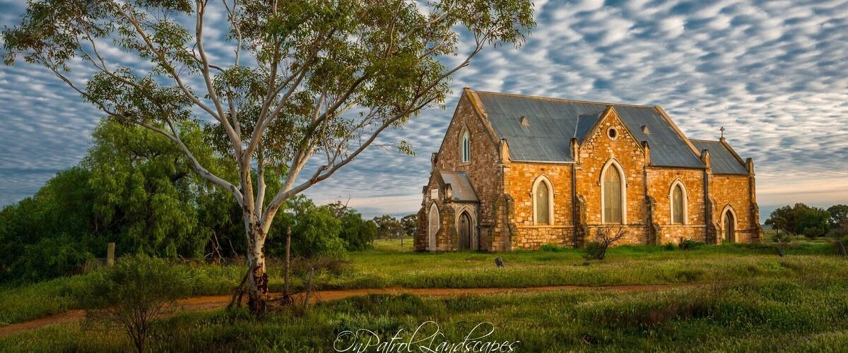 A stone church with arched windows and a large tree in front.