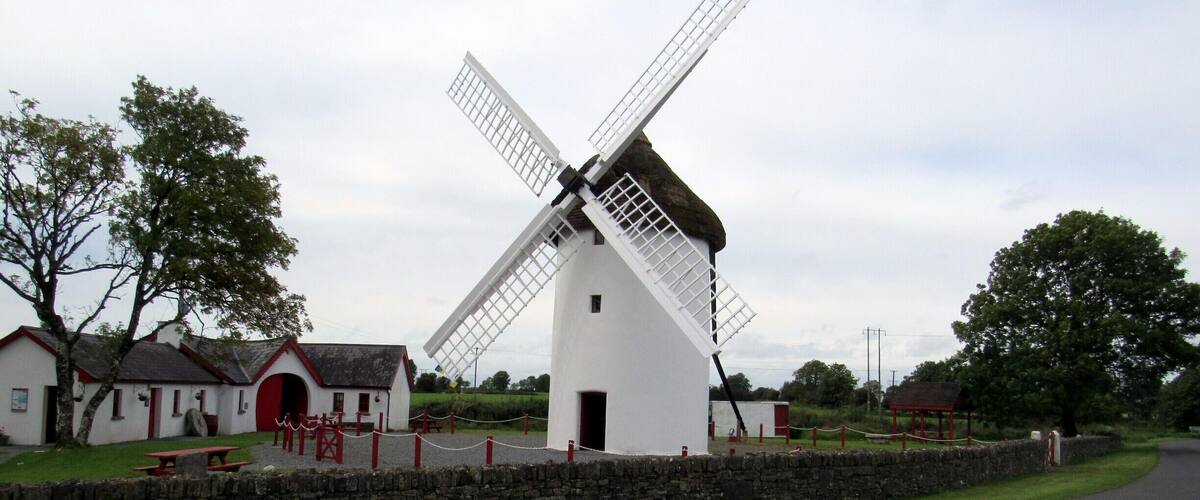 A traditional windmill with a white body and black roof, surrounded by stone walls and a grassy area with a picnic table.