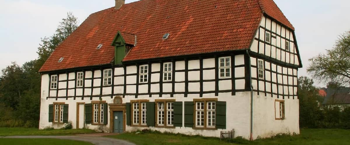 A traditional half-timbered house with a red-tiled roof, surrounded by a grassy area and a gravel path.
