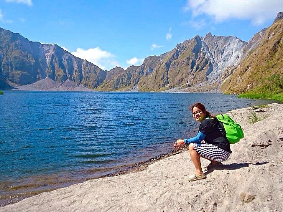 Una persona sentada en una orilla rocosa junto a un lago, con montañas al fondo.