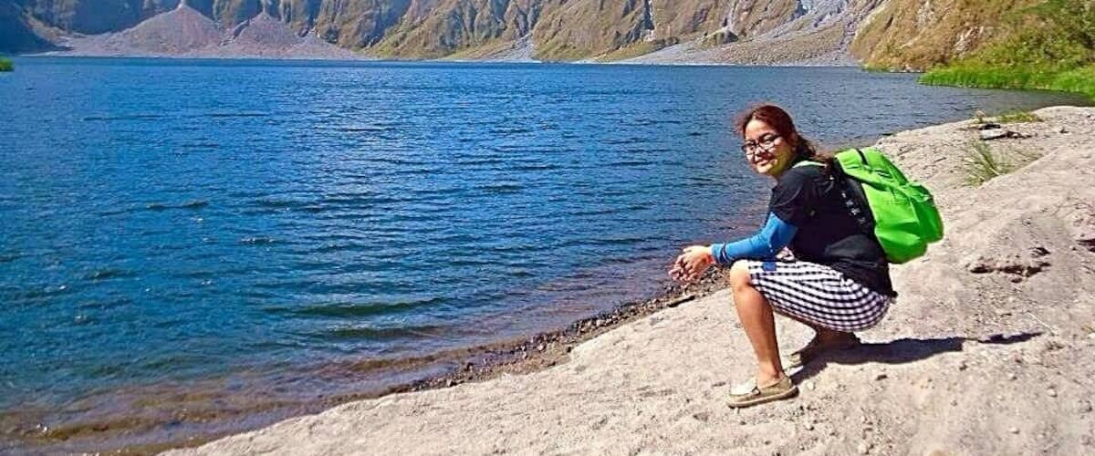 A person sitting on a rocky shore by a lake with mountains in the background.