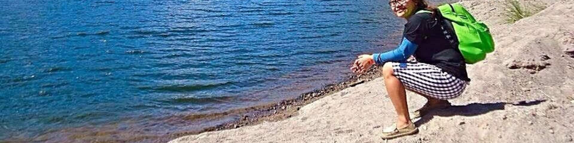 A person sitting on a rocky shore by a lake with mountains in the background.