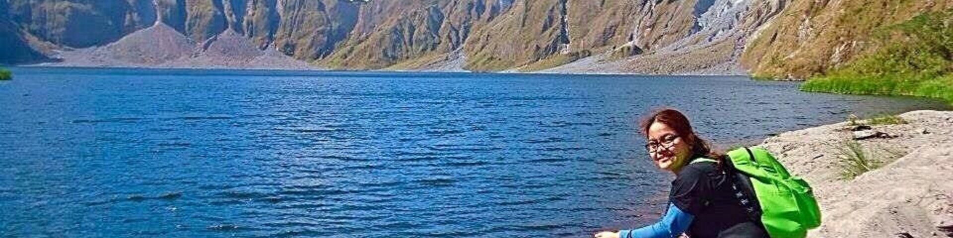 Una persona sentada en una orilla rocosa junto a un lago, con montañas al fondo.