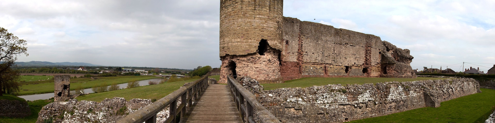 A historic stone tower with a wooden walkway leading to it, surrounded by grassy fields and a river.