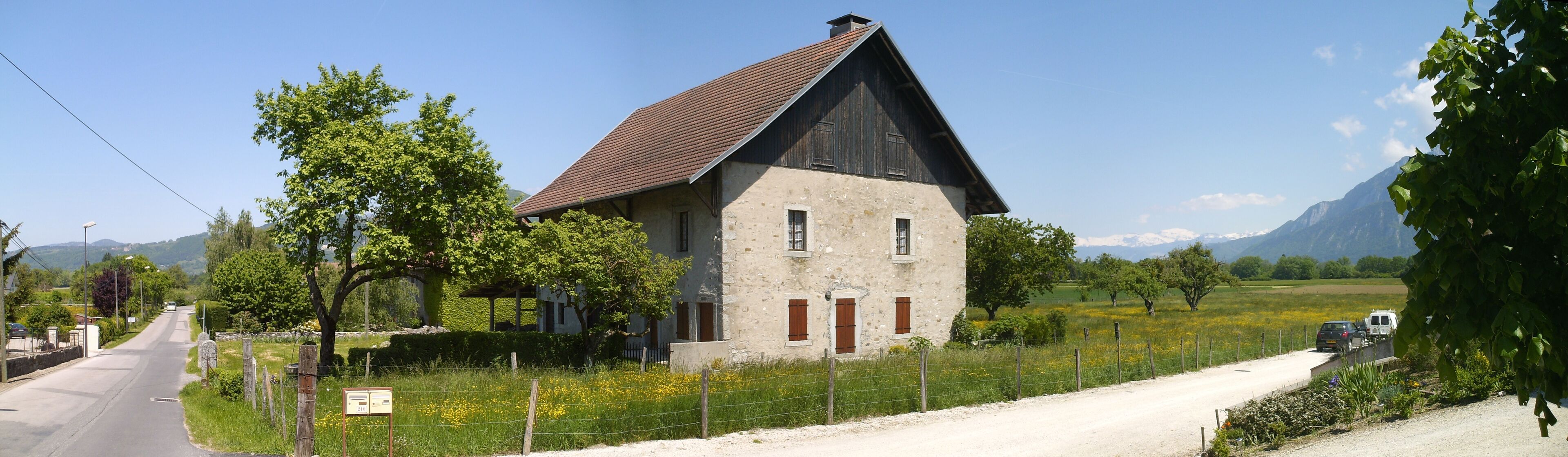 A rural house with a sloped roof, surrounded by a fence, trees, and a road.