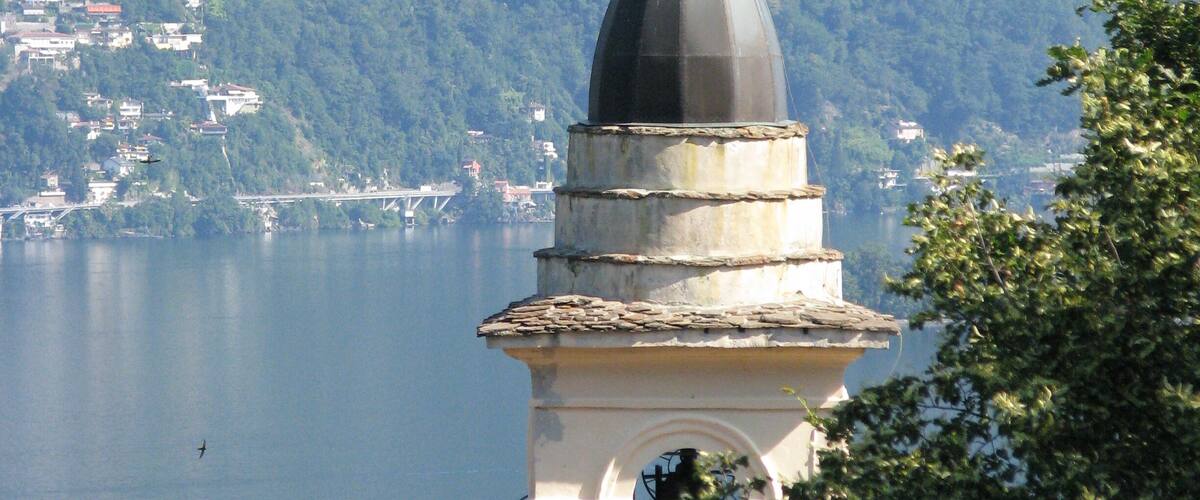 A tower with a dome and flag, overlooking a lake with a mountainous backdrop.