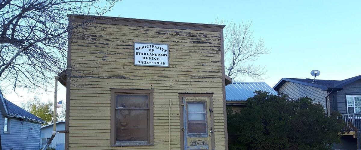 A small, weathered building labeled as the Municipality of Starland #307 Office, dating from 1920 to 1943.