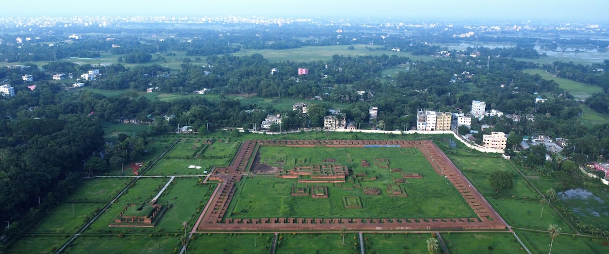 An aerial view of a large, rectangular archaeological site with multiple mounds and pathways, surrounded by lush green fields and distant urban buildings.