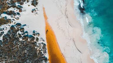 Vista aérea de uma praia com areia branca, uma faixa estreita de areia laranja e ondas oceânicas azul-turquesa.