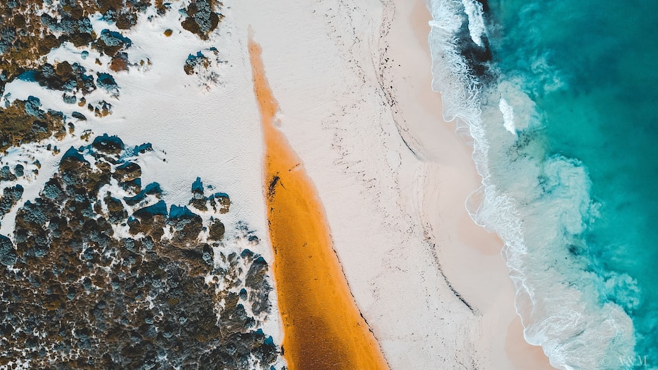 An aerial view of a beach with white sand, a narrow strip of orange sand, and turquoise ocean waves.