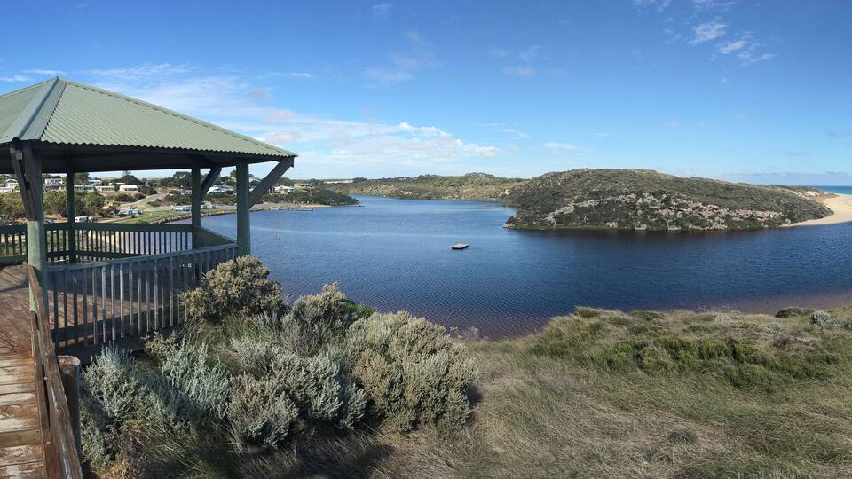 A coastal landscape with a wooden deck, a green-roofed pavilion, a sandy beach, and a small island.